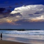 Storm clouds and dust can be seen in the sky above as people walk at dusk along Mollymook Beach